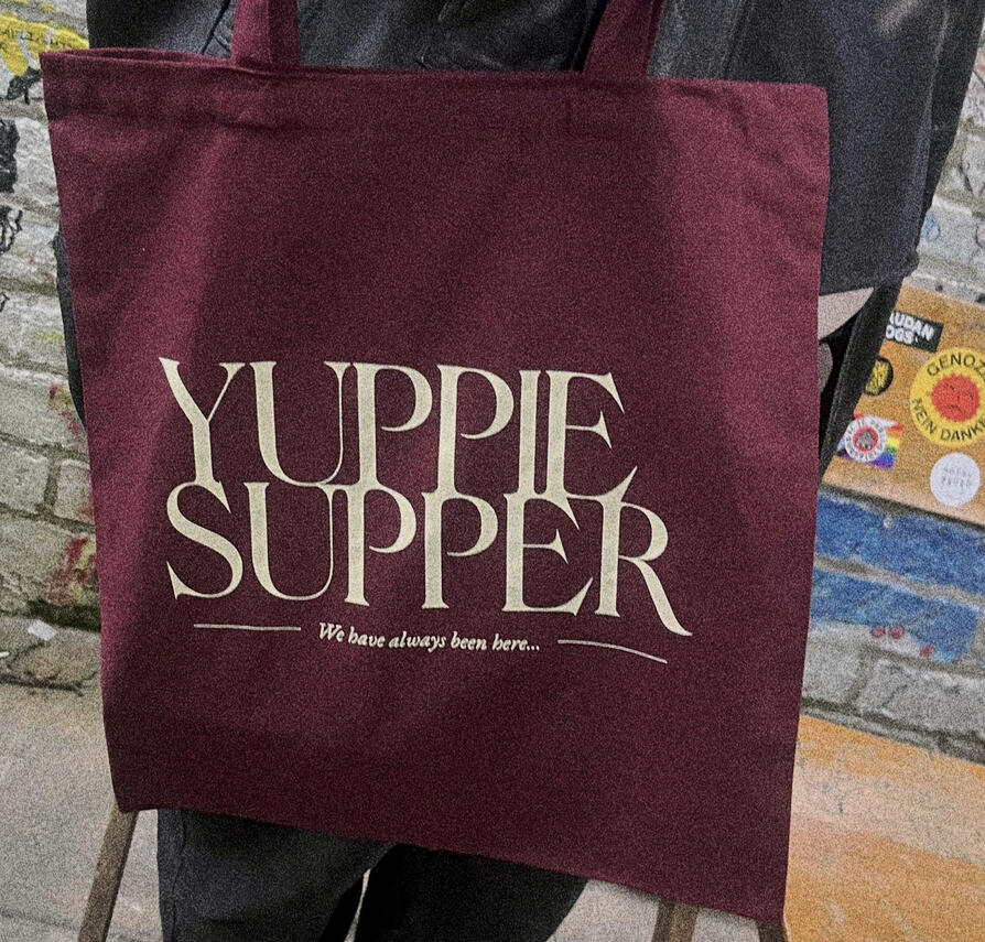 A burgundy tote bag with the Yuppie Supper logo in the centre and above the text "We have always been here..."