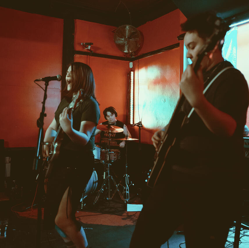 Left to right: Riley, a white woman with a brunette bob playing guitar and singing centre stage. Morgan, a white androgynous person with short, shaggy brown hair concentrating on drumming. Asher, an Irish-Chinese man with short black hair playing bass.